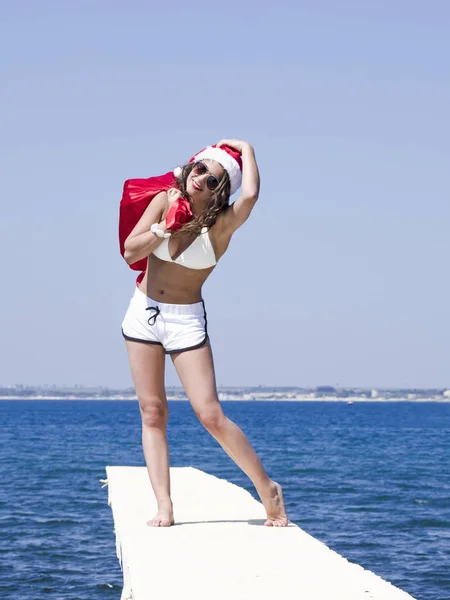 サンタさんの帽子とクリスマスのお祝いの時にプレゼントの袋と桟橋で水着立っている女性 — ストック写真