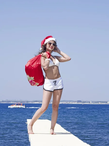サンタさんの帽子とクリスマスのお祝いの時にプレゼントの袋と桟橋で水着立っている女性 — ストック写真