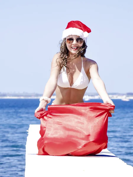 サンタ クロースの帽子とクリスマスのお祝いの時にプレゼントの袋と桟橋に座っている水着の女性 — ストック写真