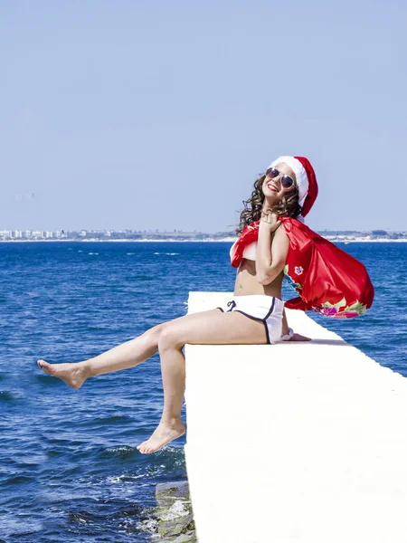 Жінка в капелюсі Санти і купальник сидить на пірсі з мішком подарунків під час святкування Різдва. Синій фон з пробілом для тексту або зображення — стокове фото