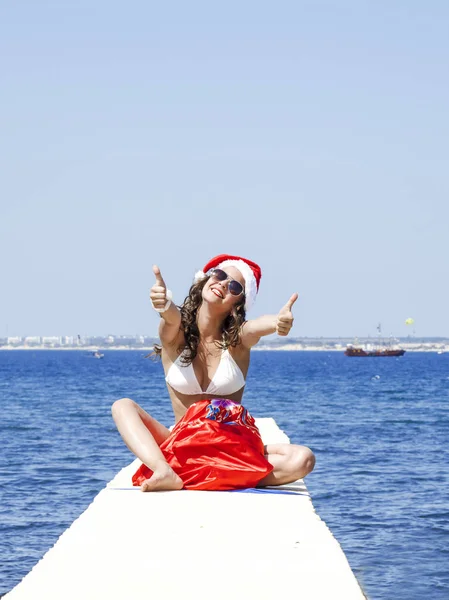 女性在圣诞老人的帽子和游泳衣坐在码头在庆祝圣诞节期间制作"Ok"手势。女孩带了一袋礼物. — 图库照片