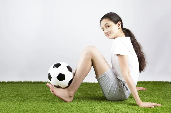 A beautiful happy girl in a sport T-shirt is holding a soccer ball on her legs. Gray background — Stock Photo, Image