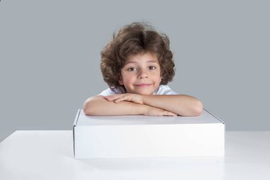 Başı beyaz kutunun üzerinde dinlenen sevimli çocuk gülümsüyor. Bir masada oturur ve kameraya bakar. Gri arka plan. Yakın plan..