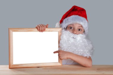 Genç Noel Baba boş bir çerçeve tutuyor ve parmağında sergiliyor. Bir masada oturur ve kameraya bakar. Yakın plan. Gri arkaplan.