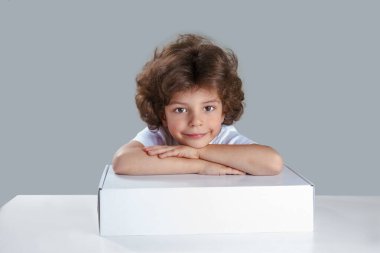 Elleri beyaz bir kutunun üzerinde katlanmış bir masada oturan tatlı kıvırcık çocuk. Kameraya bakıyor. Gri arka plan. Yakın plan..