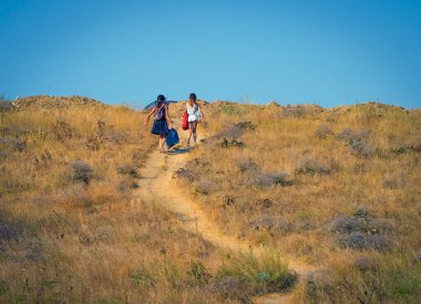 Ağır çantalı iki kız tepeye tırmanıyor. Deniz kıyısında sıcak bir yaz gününde plajdan otoparka yürüyen insanlar. Zorlukların üstesinden geliniyor.