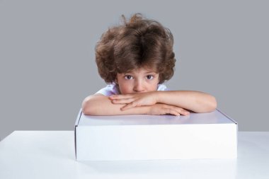 Küçük çocuk başını katlanmış elleriyle beyaz kutunun üzerine koydu. Masada oturuyor, kameraya bakıyor. Gri arka plan. Yakın plan..