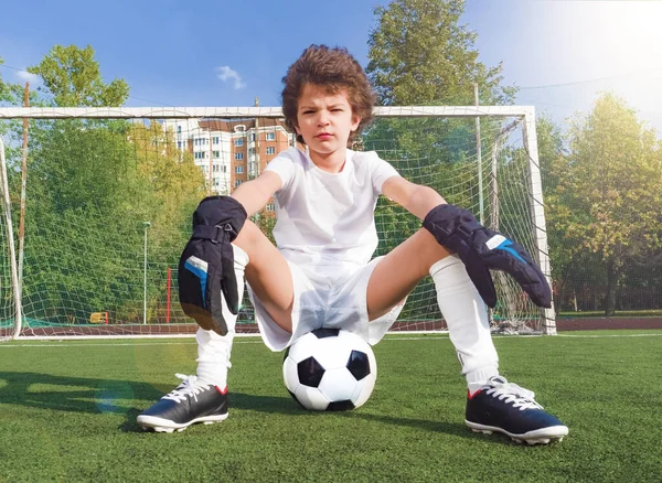 Niño pensativo sentado en la pelota de fútbol contra el gol en un campo de deporte.Niño portero en ropa deportiva de fútbol en el estadio con ball.Pose del ganador.Confianza y la voluntad de ganar concepto . —  Fotos de Stock