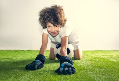 Çocuklar için futbol dersleri. Futbol topu olan sevimli, tüylü bir çocuğun portresi çimenli stüdyo arka planında yatıyor.