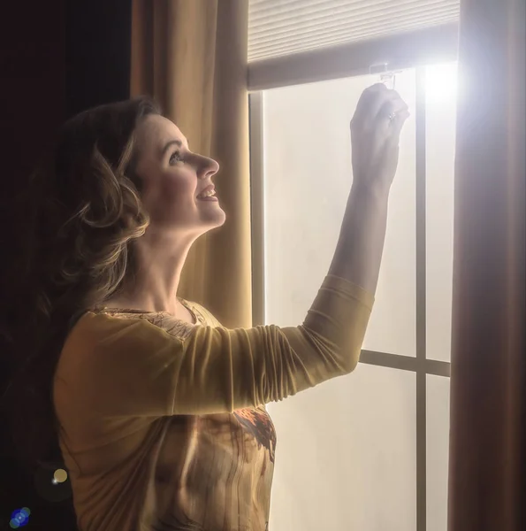 Home blinds window shades woman opening shade blind during sunny morning. Mature woman holding modern cordless top down luxury curtains indoors.