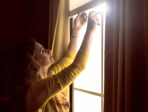 Home blinds window shades woman opening shade blind during sunny morning. Mature woman holding modern cordless top down luxury curtains indoors.