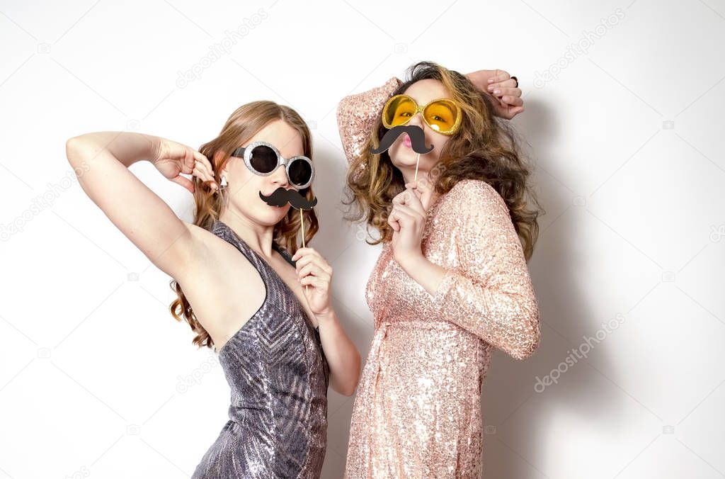 Close up portrait of two crazy funny cheerful comic attractive beautiful girlfriends wearing shiny dresses and having fun using fake mustache, isolated on light background