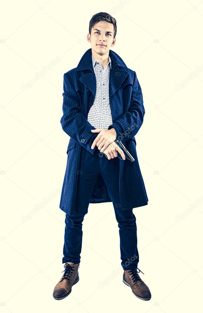 Mysterious undercover young man-gangster holding a gun and wearing a dark coat isolated on light background