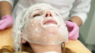 Kozmetik kliniğinde kanepede uzanmış anestezi maskeli bir kadın. Kozmetik uzmanı, kadının yüzündeki nemlendirici maskenin üstüne plastik bir film koyar. 4K