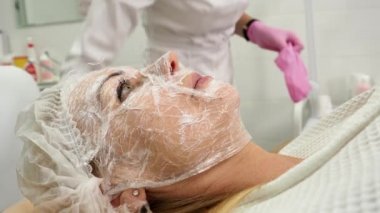 Kozmetik kliniğinde kanepede uzanmış anestezi maskeli bir kadın. Kozmetik uzmanı, kadının yüzündeki nemlendirici maskenin üstüne plastik bir film koyar. 4K