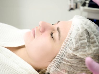 Beyaz koruyucu şapkalı ve beyaz havlulu bir kadın tıbbi bir kanepede uzanıyor ve estetik ameliyat için bekliyor..