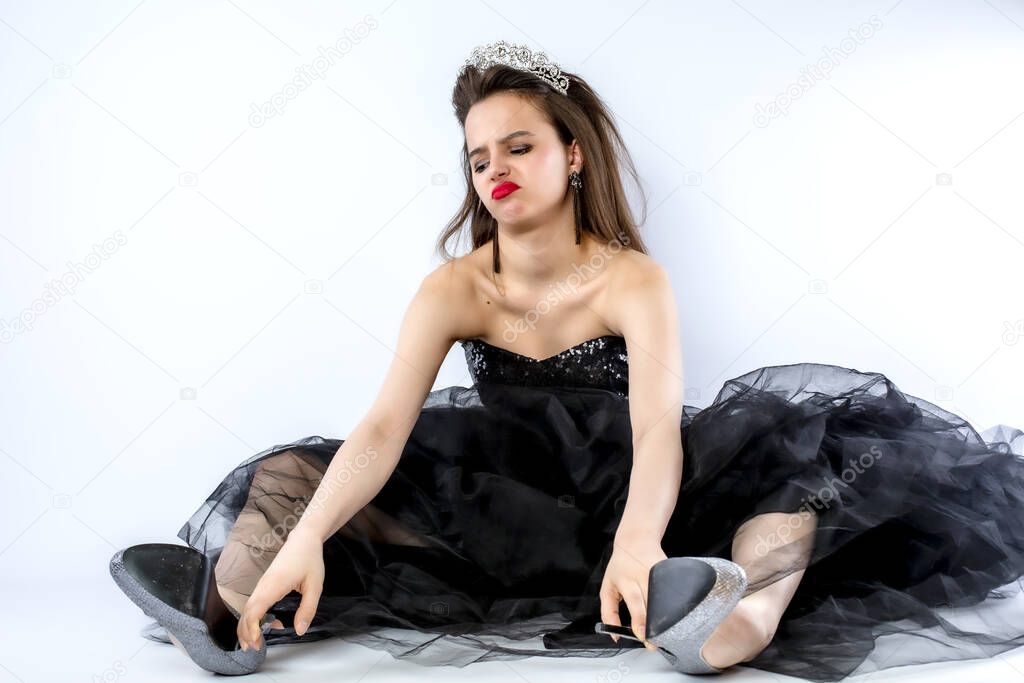 Beautiful brunette adult girl in black, long dress wearing a dadema with white stones crown sitting on the floor in a bad mood looking to the side, isolate on gray, copy space.
