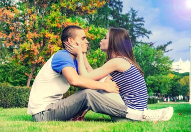 Parktaki çimlerin üzerinde oturan mutlu genç çift. Bir yaz parkında yeşil çimlerde oturan güzel genç bir çift. Kız ve erkek arkadaşı birbirlerini öpmek için uzanıyorlar. Parkta yürü