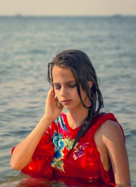 Sıcak bir günde, okyanusun turkuaz suyunda uzanan kırmızı plaj elbiseli, uzun saçlı genç esmer. Güzel üzgün kadın tatilde denizde poz veriyor..