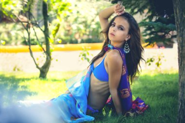 Mavi bikinili sıska bir genç kız ve boynunda mavi bir eşarp, deniz kenarında bir otelin çimlerinde oturuyor.