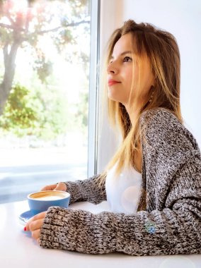 Güzel genç bayan pencerenin kenarında oturarak kahve içer. Güzel genç pencerenin kenarında oturmuş sıcak bir şeyler içiyor..