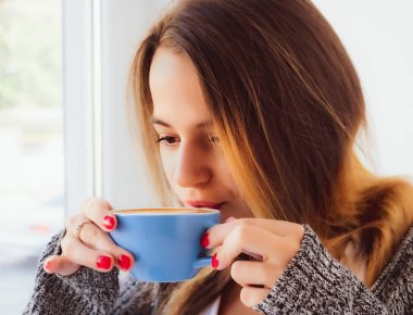 Güzel genç bayan pencerenin kenarında oturarak kahve içer. Güzel genç pencerenin kenarında oturmuş sıcak bir şeyler içiyor..