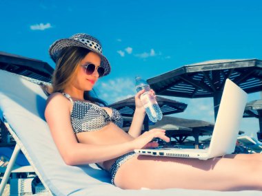 Genç Kadın Freelancer, dizüstü bilgisayar kullanarak okyanus arka planındaki sandalyede rahatça oturuyor. Sahilde. Kız Freelancer çalışıyor. Ofis dışında çalışmak. Genç bayan soğuk su içiyor..