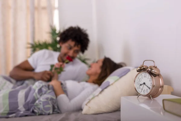 Happy young couple waking up in the morning in bed with a rose focus on clock