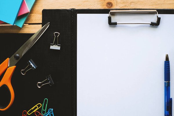 White paper on clipboard with office tool stationery (pen, scissors,notepad, binder and paper clips) on a wooden table background