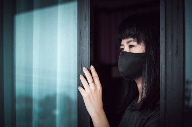 Siyah bir maske takan Asyalı kadın, kapıda dalgın duruyor ve Coronavirus veya Covid-2019 salgın konsepti ile kişisel karantina ve sosyal uzaklık için evde kalıyor.
