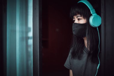 Siyah bir maske ve kulaklık takan Asyalı kadın, kapıda dalgın duruyor ve Coronavirus veya Covid-2019 salgınında kişisel karantina ve sosyal mesafe için evde kalıyor.
