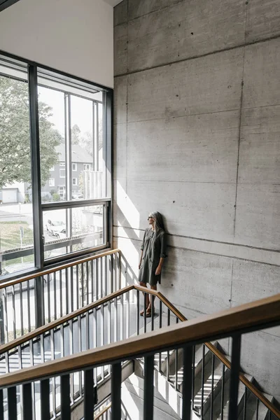 Woman with long grey hair looking out of window on staircase — Stock Photo