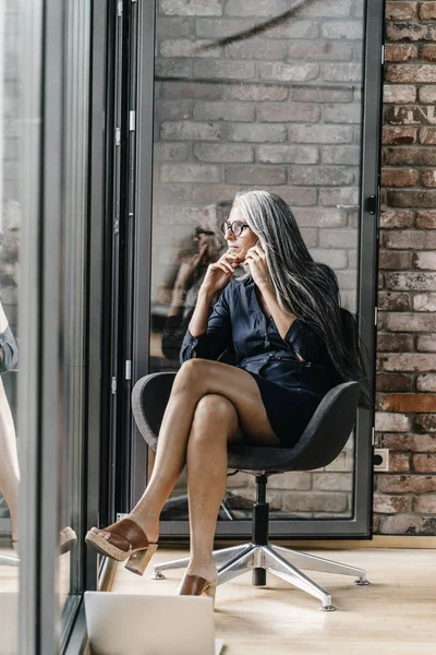 Femme avec de longs cheveux gris sur le téléphone portable assis à la fenêtre — Photo de stock