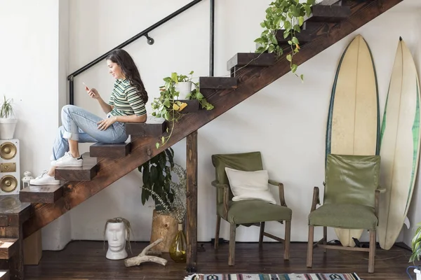 Mujer joven sentada en las escaleras en un loft mirando el teléfono celular - foto de stock