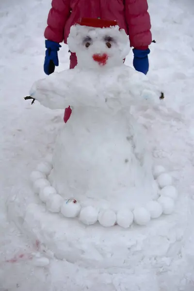 A little girl in a red overall makes a snowman from the snow. Ga — Stock Photo, Image