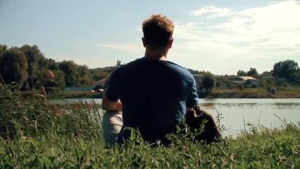 Высокий белый блондин сильный мужчина сидит на зеленой траве над озером и наблюдая прекрасный вид новые уникальные качества люди путешествуют видео — стоковое видео