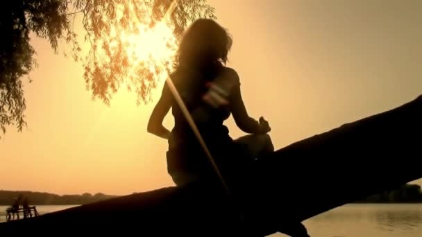 Joven de pelo largo mujer oriental sentado en el árbol al atardecer en el lago de baile observando el cielo - nueva calidad única alegre chica gente video metraje — Vídeos de Stock