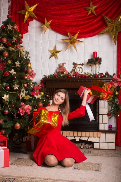 Gyönyörű lány, egy piros ruhás közelében karácsonyfa téli újévi ajándékok — Stock Fotó