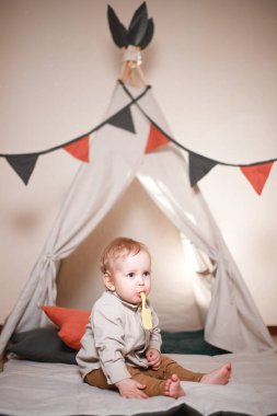 Bir çocuk, bir oğlan, evin içinde bir çadırda oturuyor ve ahşap bir arka planda oynuyor. Pantolon ve bej renkli bir gömlek giymiş.