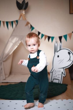 Çadırda oynayan, koyu yeşil tulum ve beyaz gömlek giyen sevimli bir çocuk.