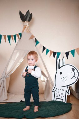 Çadırda oynayan, koyu yeşil tulum ve beyaz gömlek giyen sevimli bir çocuk.