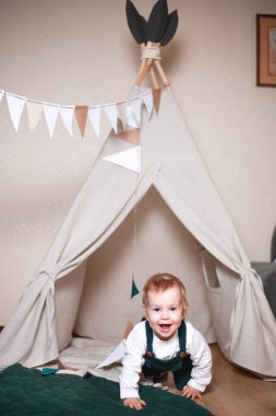 Çadırda oynayan, koyu yeşil tulum ve beyaz gömlek giyen sevimli bir çocuk.