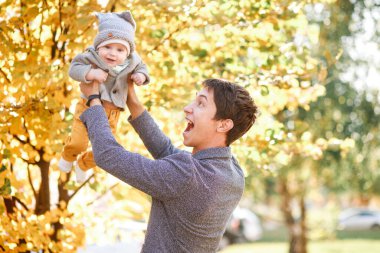 Küçük bir çocuk sonbahar manzarasının arka planında babasının kollarında oturuyor.