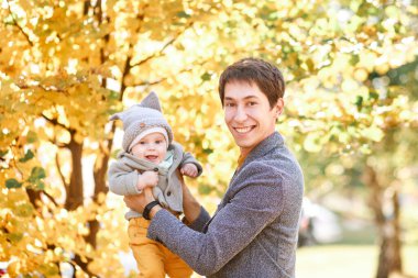 Küçük bir çocuk sonbahar manzarasının arka planında babasının kollarında oturuyor.