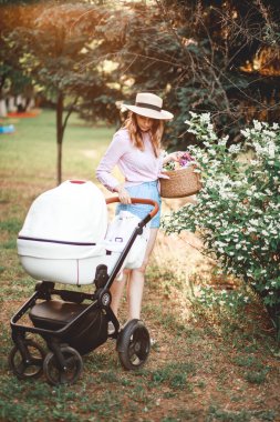 Bir kadın gün batımında parktaki yolda bebek arabasıyla yürüyor. Bebek arabasındaki bebeğe doğru eğildi.