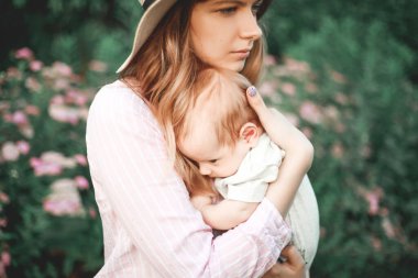 Çiçek açan bir bahçede, kollarında çocuğu olan genç bir anne. Çiçek açan ağaçların arasında duruyor..