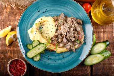 Gulaş, ahşap bir masa ve kumaş üzerinde patates püresi ve sebzelerle servis edilir.