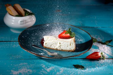 Masanın arkasında mavi bir tabakta çilekli cheesecake.
