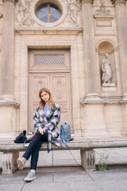 Louvre 'un yanındaki bankta oturan güzel paltolu bir kız. Fransa ve tarihi binaları ziyaret etme kavramı.