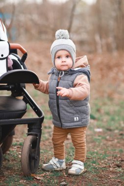 Bir yaşındaki bir çocuk pistte ilk adımlarını atıyor. Gri bir yelek ve bej pantolon giymiş seçici bir odak noktası var.